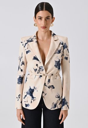 Blazer floreale in crema con motivi ad acquerello blu navy, chiusura con un solo bottone, vestibilità su misura e maniche lunghe. Abbinato a jeans scuri.