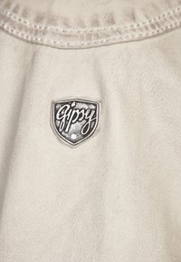 Beige leren stof met een gladde textuur, voorzien van een zilveren schildlogo met het woord "Gipsy" in een gestileerd lettertype.