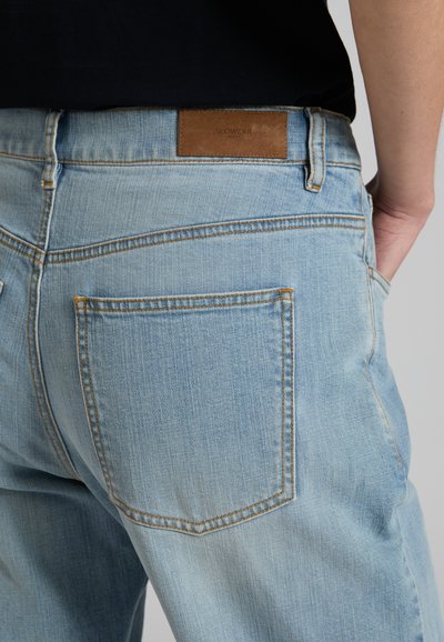 Jeans di denim azzurro chiaro con tasca posteriore e patch in pelle marrone indossati da una persona con una camicia nera.