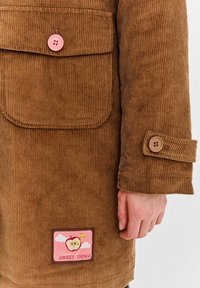 Brązowa kurtka z korduroy z różowym zapięciem na guzik, kieszenią na łatkę oraz metką z grafikiem jabłka i napisem "Sweet Things".