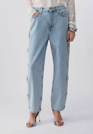 Jeans azzurri a gamba dritta con decorazioni floreali gioiello sui lati, abbinati a sandali argentati con tacco alto e una maglia bianca trasparente.