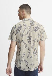 Camisa de manga corta beige con patrones florales azules y dorados, hecha de un tejido ligero, con un corte relajado y un dobladillo curvado.