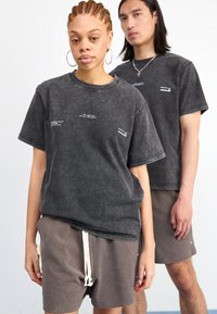 Grå, overdimensjonert bomulls-t-skjorte med kort erme, som har hvite tekstdetaljer. Modellen har på seg matchende grå shorts med snøring og knappedetaljer.