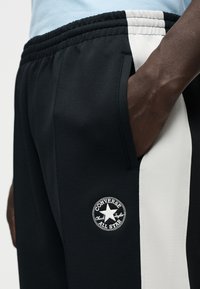 Μαύροι παντελόνες τζόγκερ με λευκή ρίγα στο πλάι και στρογγυλό σήμα Converse. Το ύφασμα φαίνεται λείο με ελαστική ζώνη στη μέση.