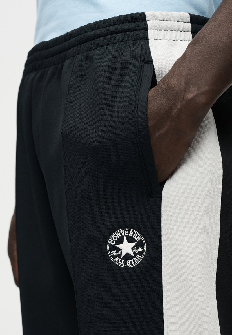 Sortte joggingbukser med en hvid stribe i siden og en rund Converse-logo patch. Stoffet ser glat ud med en elastisk talje.