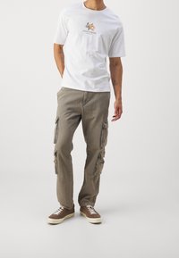 Bílý bavlněný tričko s grafikou dvou postav a nápisem "SANTORINI" vytištěným pod nimi; kombinováno s béžovými cargo kalhotami s několika kapsami a volným střihem.