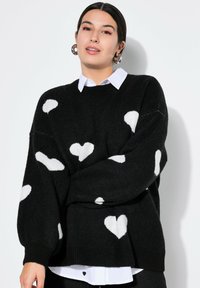 Schwarzer Pullover mit weißen Herzmustern, oversized Schnitt, gerippter Saum und langen Ärmeln, über einem weißen Hemd mit Kragen getragen. Weicher Stoff.