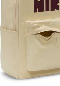Rucsac bej realizat dintr-un material durabil, cu o buzunar frontal cu fermoar și un logo maroniu îndrăzneț. Textură netedă și formă rectangulară.