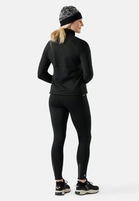 Musta urheilujakku, jossa on kaulus, yhdistettynä mustiin legginsseihin, joissa on vetoketj yksityiskohtia, yhdessä kuviollisen pipon ja urheilujalkineiden kanssa.