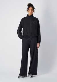 Veste coupe-vent noire avec col haut, fermeture éclair et logo ; associée à un pantalon noir à jambes larges avec des motifs sur les côtés. Texture de tissu douce.
