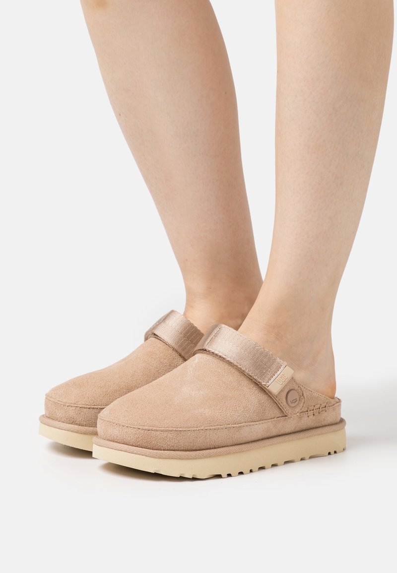 Pieds portant des sabots beige en daim avec bride ajustable et semelle en caoutchouc sur fond blanc.