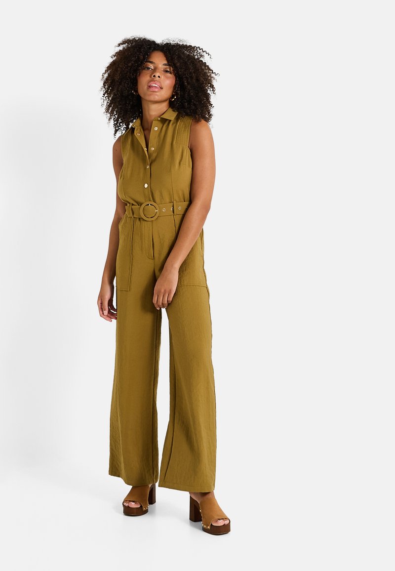 Combinaison vert olive avec un devant à boutons, taille cintrée, ceinture, jambes larges et tissu texturé ; associée à des chaussures à talons bruns.