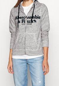 Person iført en grå zip-up hættetrøje med sorte snore og teksten "Abercrombie & Fitch New York" over en hvid skjorte og lyseblå jeans.