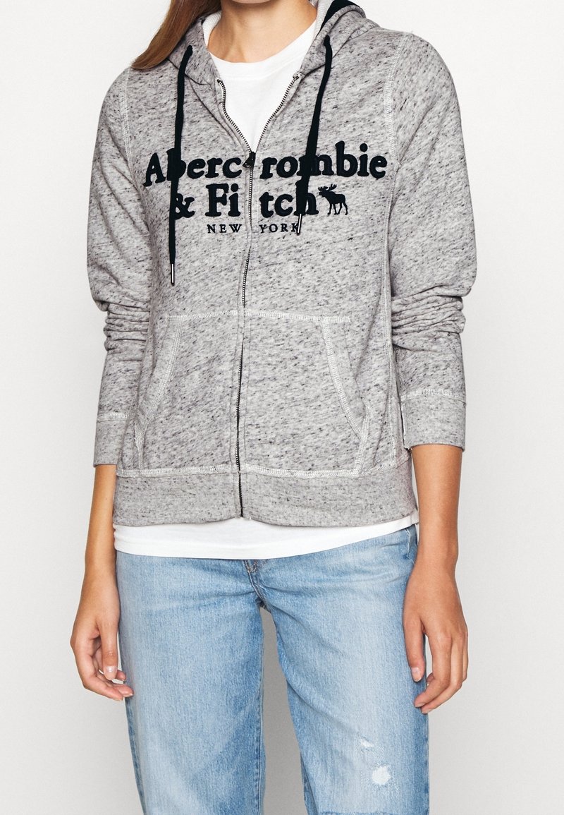 Person iført en grå zip-up hættetrøje med sorte snore og teksten "Abercrombie & Fitch New York" over en hvid skjorte og lyseblå jeans.