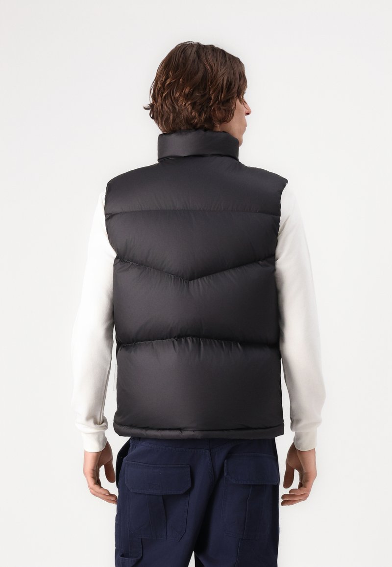 Doudoune noire sans manches avec un col haut, présentant un rembourrage matelassé en motif chevron. Portée sur une chemise à manches longues blanche, la personne tournant le dos.