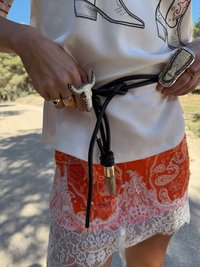 Cinto de couro preto com detalhes prateados, usado sobre uma saia laranja com padrão paisley e acabamento em renda branca. Vários anéis visíveis nos dedos.