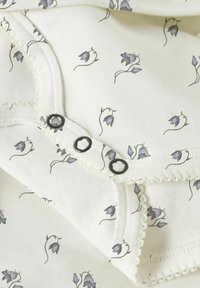 Bomullsbody med kremfarget bakgrunn og grå blomster. Den har tre svarte knapper og skåret kant langs halsen.