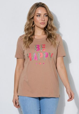 Vrouw met golvend blond haar draagt een bruin T-shirt met meerkleurige "BE HAPPY" tekst en lichtblauwe spijkerbroek, staand tegen een effen achtergrond.