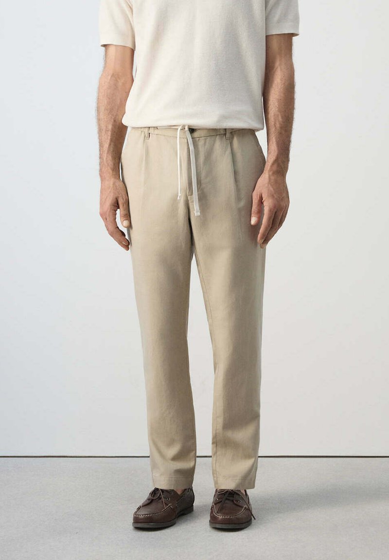 Homme portant un pantalon beige avec cordon, un t-shirt blanc et des chaussures en cuir marron à lacets, debout devant un fond uni.