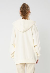 Sweat à capuche crème avec cordon de serrage, coupe ample, manches larges et texture douce. Design simple sans motifs ou accents proéminents.