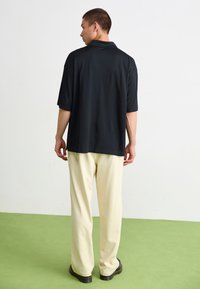 Μαύρο polo πουκάμισο κοντό μανίκι με χαλ�αρή εφαρμογή, συνδυασμένο με κρεμ παντελόνι φαρδιάς γραμμής, σε θέα από πίσω πάνω σε πράσινο δάπεδο.