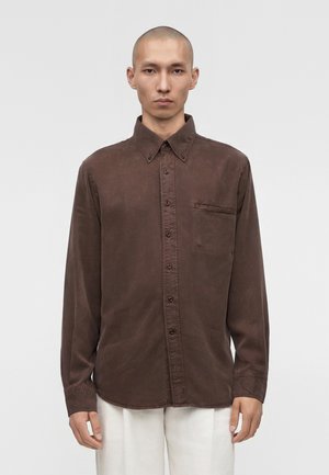 Hombre con la cabeza afeitada que lleva una camisa marrón oscuro de manga larga abotonada y pantalones blancos, de pie frente a un fondo claro y liso.