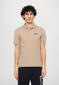Beige polo shirt with black collar and sleeve accents, featuring a black EA7 logo. Made of cotton, it has a classic fit and short sleeves.