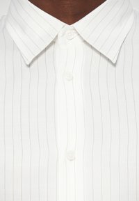 Gros plan d'une chemise blanche à col avec de fines rayures verticales noires et des boutons blancs, portée par une personne à la peau foncée.