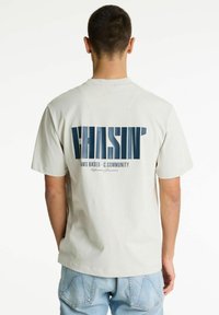 Camiseta de manga corta gris claro con gran gráfico en azul marino "CHASIN'" en la parte posterior, con texto adicional abajo. Tejido suave de algodón.