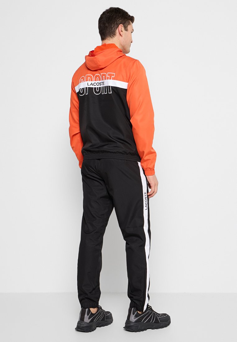 Un homme portant un vêtement de sport Lacoste noir et orange avec une veste à capuche et un pantalon présentant des rayures latérales blanches et des logos, se tenant contre un fond blanc.