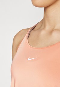 Pfirsichfarbenes Sporttanktop aus glattem, matter Stoff. Mit einem Rundhalsausschnitt und einem kleinen weißen Nike-Logo auf der Brust.