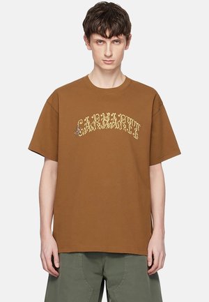 Giovane uomo che indossa una t-shirt Carhartt marrone con logo giallo arcuato e pantaloni verde oliva, in piedi su uno sfondo semplice.