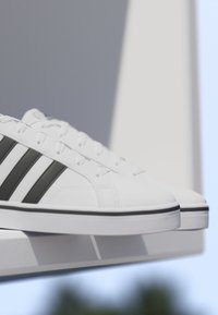 Zapatillas blancas con rayas negras sobre una estantería blanca, fondo desenfocado con contraste de luz y sombra.