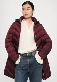 Vrouw in een bordeauxrode pufferjack over een crèmekleurige geribbelde trui, met haar handen in de zakken van hoge taille blauwe jeans, staande tegen een effen achtergrond.