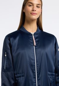 Navy blauwe bomberjack met satijnen afwerking, een geribbelde kraag, een voorkeursrits en zijzakken; heeft een ritszak op de mouw.