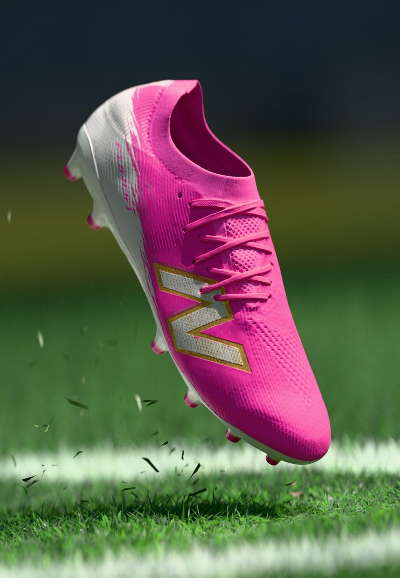 Scarpetta da calcio rosa brillante con logo "N" bianco e oro che solleva erba su un campo verde con linee bianche tracciate.