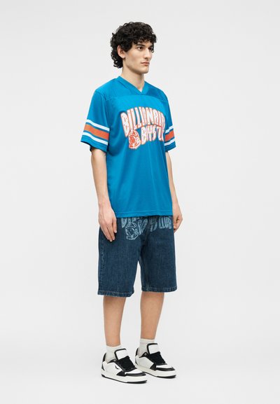 Νέος άνδρας που φοράει μπλε αθλητική φανέλα "Billionaire Boys Club", σκούρα τζιν σορτς με εκτύπωση λογότυπου, λευκές κάλτσες και ασπρόμαυρα αθλητικά παπούτσια.