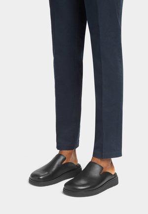 Schwarze Slipper bestehen aus glattem Leder mit einer runden Zehenpartie, einer flachen Sohle und einem offenen, geschwungenen Design an den Seiten.