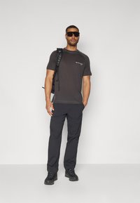 Dunkelgraues sportliches T-Shirt mit kurzen Ärmeln, kombiniert mit schwarzen Wanderhosen und robusten schwarzen Schuhen. Zubehör sind ein Rucksack und eine Sonnenbrille.