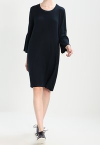 Tmavé pletené šaty mají kulatý výstřih, dlouhé zvonové rukávy a volný střih. Skvěle se hodí k černým teniskám na šněrování pro ležérní vzhled.