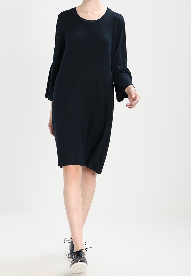 Tmavé pletené šaty mají kulatý výstřih, dlouhé zvonové rukávy a volný střih. Skvěle se hodí k černým teniskám na šněrování pro ležérní vzhled.