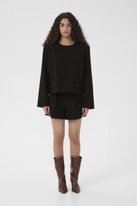 Kobieta o długich, kręconych włosach, ubrana w czarną, długorękawową bluzkę w cienkie paski i pasujące krótkie spodenki, do tego brązowe skórzane buty za kostkę, stojąca.