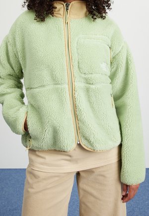Veste polaire vert clair avec bordure beige, fermeture éclair à l'avant et poche poitrine, portée avec un pantalon beige, main du mannequin dans la poche.