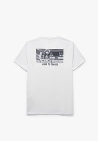 Camiseta de algodón blanca con un gráfico trasero de un atleta saltando sobre un obstáculo, con el texto negro "SALTA AL OBJETIVO" debajo de la imagen.