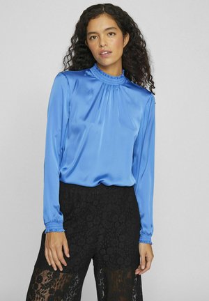 Blouse en satin bleu avec un col froncé et des manches longues, associée à un pantalon en dentelle noire avec des motifs floraux et des sections transparentes.