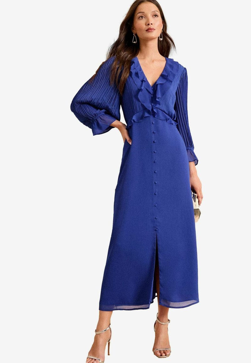 Abito blu di media lunghezza con scollo arricciato, maniche lunghe plissettate, chiusura frontale con bottoni e spacco sull'orlo. Tessuto con texture liscia.