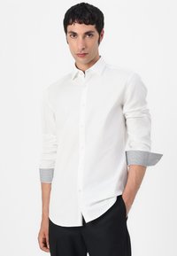 Cămașă albă cu nasturi, cu mâneci lungi, având accente cu model la manșete. Material moale; croială ajustată cu hem rotunjit. Pantaloni negri dedesubt.