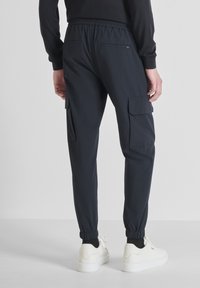 Persona che indossa pantaloni cargo neri con polsini elasticizzati e sneakers bianche, in piedi contro uno sfondo chiaro e semplice.