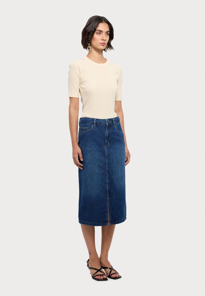 Top bej din material elastic, cu mâneci scurte, asortat cu o fustă din denim albastră, până la genunchi, care are o fanta în față și detalii simple cu nasturi metalici.