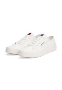 Baskets en toile blanches avec une semelle en caoutchouc texturé, présentant un laçage à l'avant et un patch logo sur la languette. Design minimaliste.